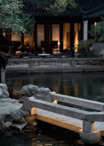 A serene garden scene featuring a stone bridge over water, with a traditional Chinese building in the background.