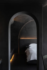 A minimalist bedroom with a concrete headboard and floor, featuring a single white bed and soft lighting.