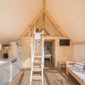 A small cabin interior with wooden paneling and a loft space accessed via a ladder. Features a kitchenette, seating area, and