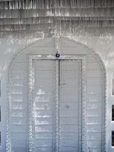 A white metal door with a decorative fringe on top, set against white walls with sunlight casting shadows.