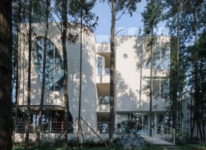 Modern residential building featuring glass walls and concrete structure surrounded by greenery.