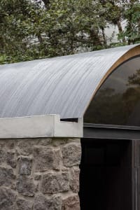 A curved metal roof with a stone wall beneath it, surrounded by greenery.