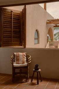Minimalist residential balcony featuring woven rattan chair, terracotta tiles, and wooden shutters.