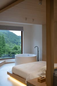 A minimalist bedroom featuring a freestanding bathtub overlooking a lush green landscape through a large window.