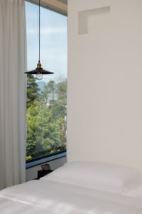 A modern white bedroom with a large window overlooking green trees and mountains, featuring a minimalist design with white be