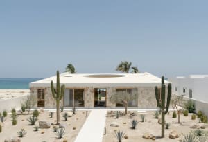 Modern building exterior with concrete walls and a circular cutout on the roof, set against a beach and ocean backdrop.