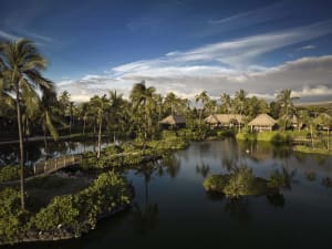A serene waterscape featuring a resort with thatched-roof huts and lush vegetation under a clear, partly cloudy sky.