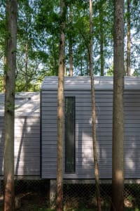 A modern residential exterior with wooden siding and a slanted roof, surrounded by trees and natural light.