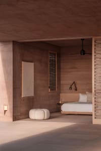 A minimalist bedroom with clay walls and a white bed, featuring a wall-mounted lamp, a window with view, and a pouf.