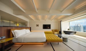 A modern hotel bedroom featuring a king-sized bed with a yellow leather headboard, a yellow leather daybed, and a large windo