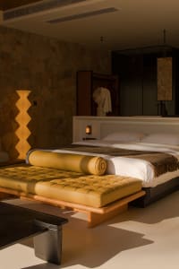 A minimalist bedroom featuring a wooden bed frame, a leather bench, and a stone wall.