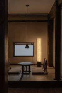 A minimalist living room with tan tones, featuring a small round table, cushioned floor seating, a tall cylindrical lamp, and
