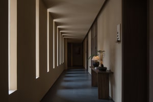 A minimalist corridor with concrete walls and floor, featuring rectangular windows and a wooden side table with a lamp and po