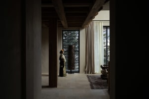 A person dressed in traditional attire standing in a minimalist architectural interior with exposed wooden beams and large wi