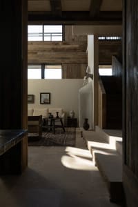 A minimalist residential interior with stone and wood materials, featuring a living area with large windows and a staircase.