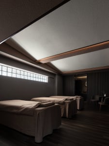 Spa room with three massage beds covered with brown fabric, dark interior, and small window with square glass panels.