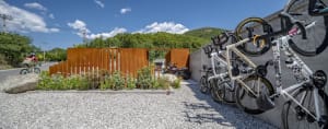 Bicycles parked on a gravel surface with a metal fence and greenery in the background.