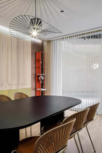 Modern dining room with a black oval table, wooden chairs with geometric patterns, glass pendant light, and red bookshelf.