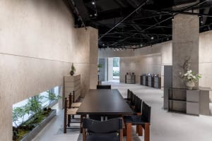 A modern restaurant interior featuring concrete walls, wooden furniture, a long dining table with black chairs, and plants.