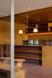 A modern kitchen with wooden cabinets, shelves, and stools, featuring a stainless steel countertop and warming lights.