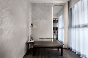 A minimalist living room with concrete walls and a small bench, featuring a modern light fixture and a large mirror.