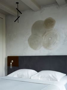 Minimalist bedroom with a textured plaster wall and a grey upholstered headboard with a brown fabric inset. The bed is neatly