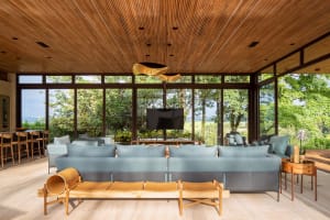 Modern living room featuring a blue sofa, wooden chairs, and a TV mounted above a table, with large windows providing a sceni