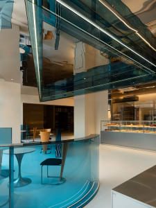 Modern restaurant interior with glass partition separating dining and kitchen areas, featuring tables, chairs, and a kitchen visible through the glass wall