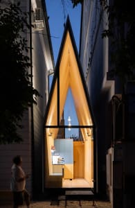 A house facade with a large wooden window featuring a unique triangular design, and a door on the bottom right corner, creating a cozy and inviting atmosphere