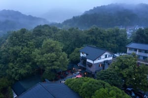 A house surrounded by trees in a forest, with a black roof and a balcony, featuring outdoor seating and a peaceful atmosphere