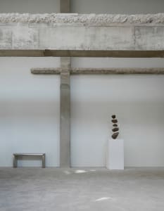 Empty industrial space with concrete walls and floors, featuring three sculptures and a bench, with a minimalistic and stark atmosphere