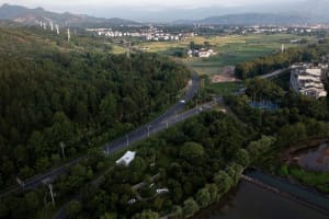 Aerial view of a residential area with houses, trees, and roads, surrounded by greenery and featuring parked cars