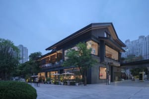 A modern building with a large storefront on the ground floor, featuring a mix of black and brown tones, with umbrellas outside and potted plants adding to the welcoming ambiance