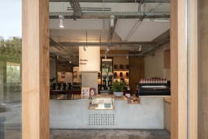 A warm and inviting coffee shop with a rustic atmosphere, featuring a counter, dining table, chairs, potted plants, and various cups and bottles on display
