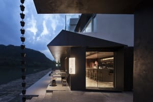 Modern restaurant with a black concrete exterior, glass door, and interior dining area with tables and chairs, situated near the water