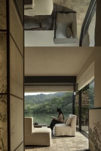 A serene living space with a modern design, featuring a woman sitting on a white chair in front of a window overlooking a lake, with a couch and another chair nearby