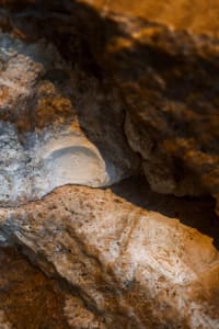 A dimly lit underground cave with a rocky wall and a small opening, lit by natural sunlight filtering through the entrance