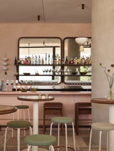 A pink-themed bar area with a long counter, wine glasses, and chairs, surrounded by bottles and conveying a cozy atmosphere