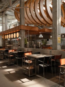 Industrial kitchen with a modern dining area, featuring a large dining table, appliances, and potted plants, in a clean and organized space