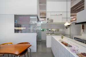 A modern kitchen with white walls, tile floors, and a dining table with a bowl and two chairs, surrounded by various kitchen items and potted plants, in a brightly lit and inviting space