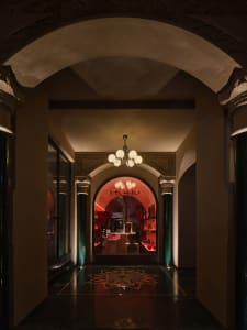 Ornate hallway with red walls, a grand archway, and chandeliers providing illumination