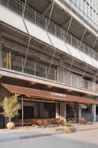 A restaurant patio area with a concrete facade, featuring multiple chairs and dining tables arranged for comfortable seating, surrounded by a modern building exterior