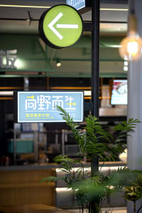 A well-lit Asian restaurant with a green sign featuring Chinese characters, surrounded by potted plants and two TVs on the wall