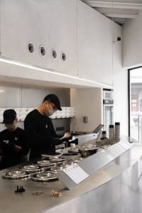 Industrial kitchen with two people working, wearing face masks, surrounded by appliances, bowls, and cups