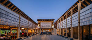 An open-air restaurant with a wooden structure and thatched roof, featuring a dining area with chairs and tables in a tropical setting