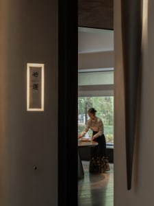 A woman stands near a dining table with a book, surrounded by a room with a doorway and a potted plant