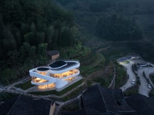 A serene photo of a modern house on a hillside, surrounded by trees and mountains, with large windows and concrete exterior walls