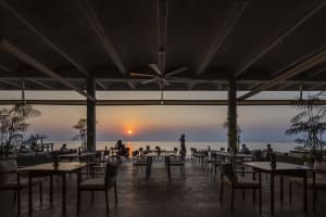 Outdoor restaurant with modern seating, concrete, wood, and metal materials, and warm lighting near a body of water
