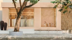 A building with a pink facade features a large window and seating options, including two benches and three chairs, in a calm and inviting atmosphere