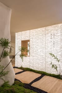 A well-lit interior space with a white wall featuring Asian writing, surrounded by potted plants and natural light from a window